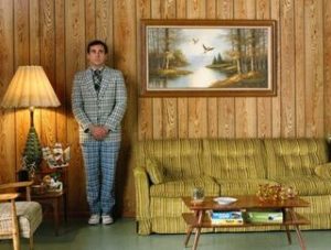 Guy in plaid suit standing against wood paneled wall in retro living room. 